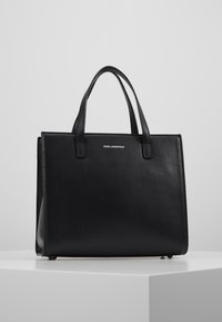 Černá kožená taška přes rameno se strukturovaným tvarem, dvojitými rukojeťmi nahoře a embosovaným logem na přední straně. Hladká textura s jemnými detaily zrnité struktury.