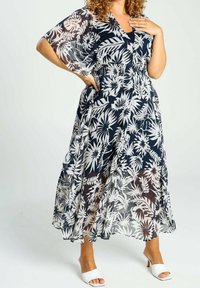 Kvinna i en genomskinlig svart och vit blommig midiklänning med ärmar till armbågen och vita klacksandaler, poserande med handen på höften och bröstet.