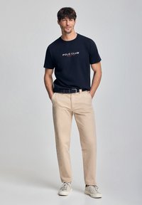 Marinblå t-shirt med "POLO CLUB"-logga, beige chinos och ljusa sneakers. Avslappnad design med lös passform och mjuk tygstruktur.