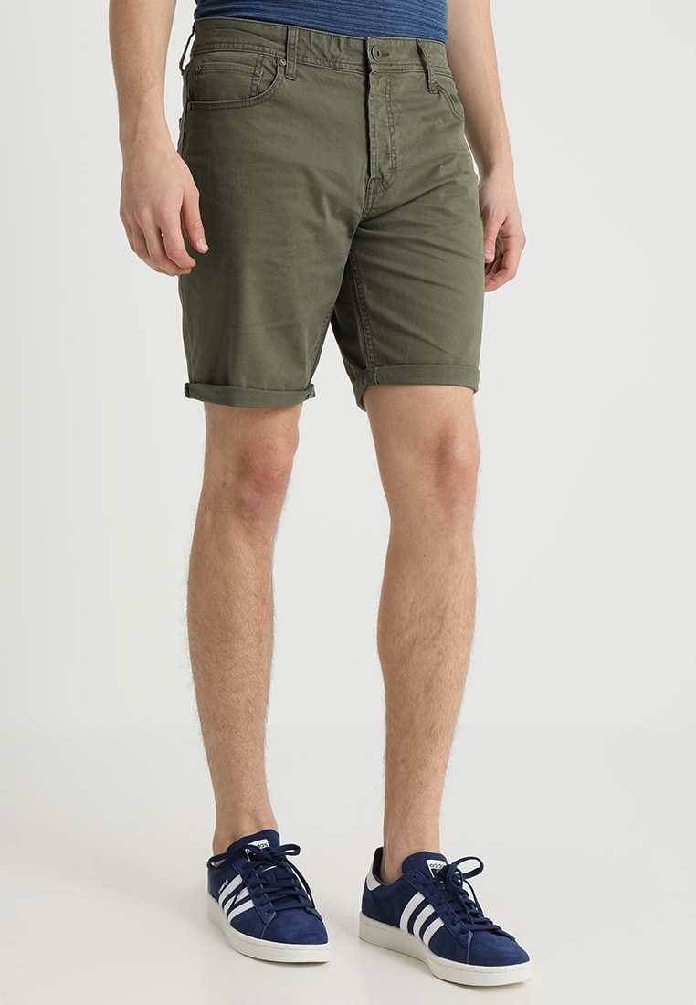 Mann in aufgerollten olivgrünen Shorts, blauen gestreiften Sneakers und einem blauen Hemd, der vor einem schlichten hellen Hintergrund steht.