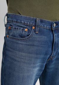 Jeans in denim blu con taglio classico, caratterizzati da rivetti in ottone, texture invecchiata e una tasca frontale visibile.