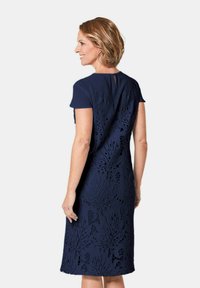 Marineblauwe jurk met korte mouwen, voorzien van een getextureerd bloemenkantontwerp en een keyhole-detail op de rug. Knie-lengte met een aansluitende silhoutte.