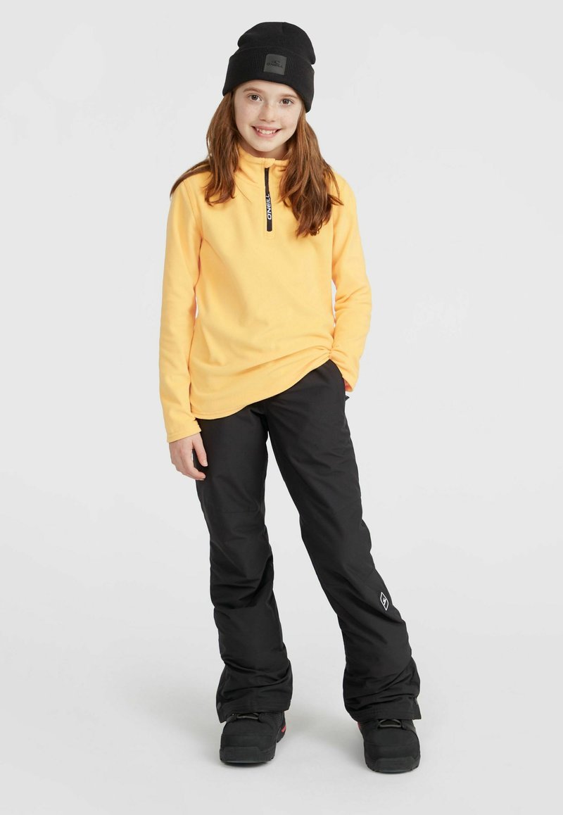 Geel fleece trui met een rits op de borst, gecombineerd met zwarte waterdichte broek en zwarte laarzen. De outfit bevat een beanie met een logo patch.