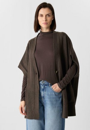 Brązowy oversize kardigan z krótkimi rękawami, wykonany z miękkiego materiału. Posiada zapięcie na guziki i prążkowane wykończenia. Nosi się go na ciemny top i dżinsy.