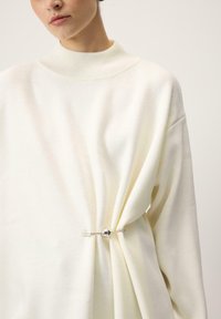 Kreemikas kudumiga kampsun kõrge kraega, millel on drapeeritud disain ja hõbedane ümmargune nõel, mis kogub ühelt küljelt kangast.