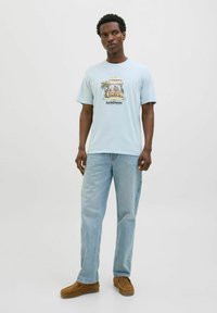 Lichtblauwe katoenen t-shirt met een grafische print van een strandscène met palmbomen. Gecombineerd met vervaagde blauwe spijkerbroeken en bruine instapschoenen.