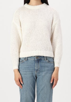 Personne portant un pull en grosse maille blanc à manches longues et un jean taille haute bleu clair, debout devant un fond uni.