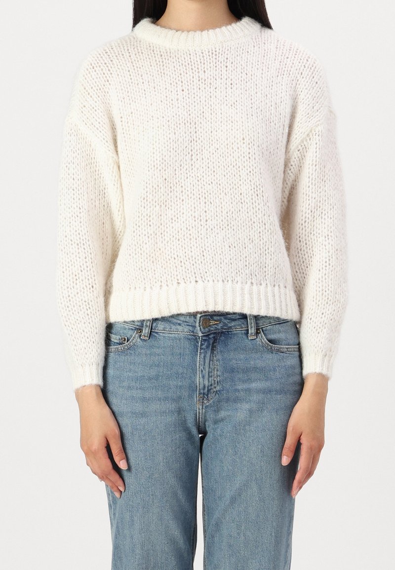 Personne portant un pull en grosse maille blanc à manches longues et un jean taille haute bleu clair, debout devant un fond uni.