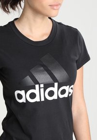 Černé bavlněné tričko s velkým bílým logem "adidas" a třemi diagonálními pruhy v gradientu šedé barvy. Krátké rukávy a kulatý výstřih.