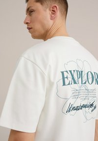 Fehér pamut póló, amelyen a hátsó oldalon egy nagy kék "EXPLORE" grafikus szöveg és virágos minta található, rövid ujjal és kerek nyakkivágással.