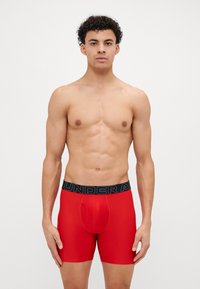 Boxeri roșii, cu o bandă elastică neagră pe care este inscripționat textul "UNDER ARMOUR". Material neted, elastic și design ajustat. Fără modele.