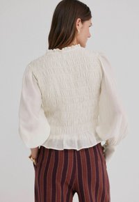 Kremowa bluzka z marszczonymi detalami i bufiastymi, przezroczystymi rękawami. Falbaniasty dekolt i dół, w połączeniu z bordowymi spodniami w paski.