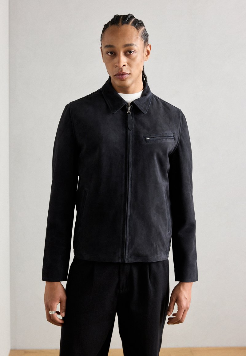 Schwarze Wildlederjacke mit strukturiertem Kragen, vorderem Reißverschluss und Brusttasche. Modell trägt sie mit passenden dunklen Hosen.
