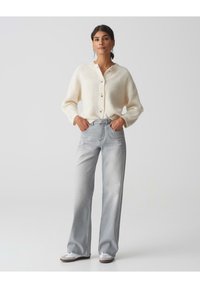 Crème gebreide cardigan met knopen, gecombineerd met lichtgrijze recht-toe-recht-aan jeans en witte sneakers. Eenvoudig, textuurontwerp met praktische styling.