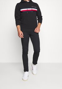 Czarna bluza z kapturem z paskiem na klatce w kolorach czerwonym, białym i niebieskim; zestawiona z dopasowanymi czarnymi spodniami i białymi sneakersami z kolorowymi akcentami.