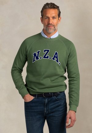 Man met kort haar en baard, gekleed in een groene trui met de letters "N.Z.A.", lichtblauw overhemd met kraag en donkerblauwe spijkerbroek, staand met een hand in de zak.