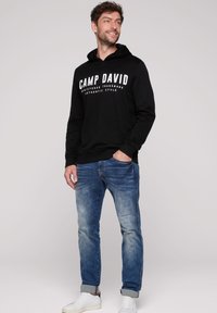 Sudadera negra con el logo "CAMP DAVID" en blanco, combinada con jeans de mezclilla azul desgastados y zapatillas blancas. El modelo se posa de manera casual con las manos en los bolsillos.