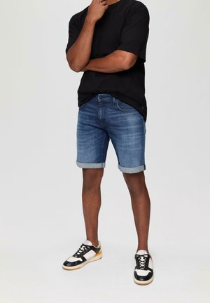 Shorts en denim bleu moyen avec des ourlets retroussés, associés à un t-shirt noir. Les chaussures sont des baskets noires et blanches avec une semelle en gomme.