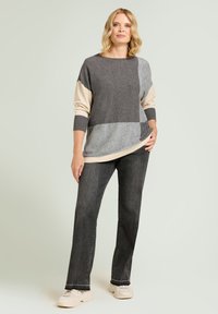 Maglione a blocchi di colore grigio e beige con collo rotondo e maniche lunghe, abbinato a jeans grigio scuro e sneakers chiare su uno sfondo chiaro.