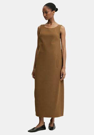 Vestito marrone senza maniche con una silhouette dritta, tasca laterale e texture liscia. Indossato con scarpe basse nere caratterizzate da un motivo strutturato.