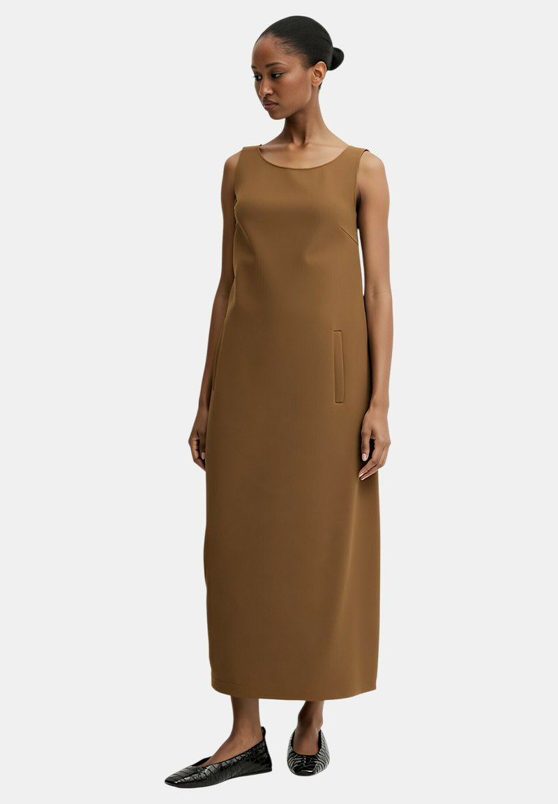 Vestito marrone senza maniche con una silhouette dritta, tasca laterale e texture liscia. Indossato con scarpe basse nere caratterizzate da un motivo strutturato.