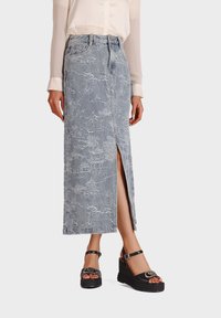 Gonna midi in denim di colore azzurro chiaro, con intricati motivi bianchi e uno spacco frontale. Abbinata a sandali con tacco a cuneo neri.