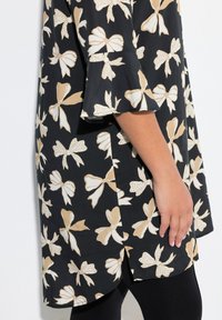 Tunic nera con grandi motivi a fiocco beige e crema, maniche a campana e orlo curvo. Tessuto liscio con una texture leggera.