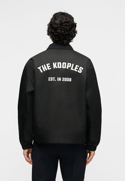 Άτομο με σγουρά σκουρόχρωμα μαλλιά φοράει μαύρο μπουφάν με λευκό κείμενο "THE KOOPLES EST. IN 2008" στην πλάτη, στέκεται μπροστά σε απλό φόντο.