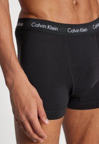 Schwarze Boxershorts aus Baumwolle mit einem dünnen schwarzen elastischen Bund, auf dem "Calvin Klein" in weißer Schrift steht. Glatte Textur, enganliegende Form.