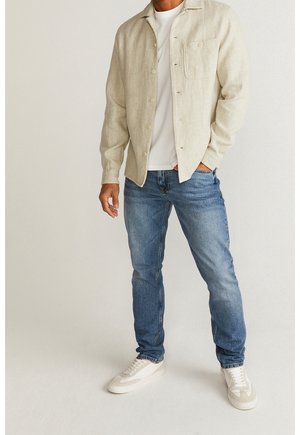 Uomo che indossa una camicia beige chiaro abbottonata sopra una t-shirt bianca, jeans blu e sneakers bianche, in piedi contro uno sfondo chiaro e semplice.