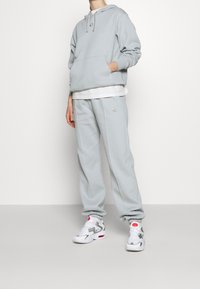 Ljusgrå hoodie med framficka och en liten logotyp, kombinerad med matchande sweatpants och vita sneakers med röda detaljer.