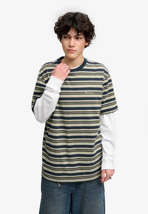 Ung person med krøllet mørkt hår iført lagdelt stribet kortærmet skjorte over hvide langærmede trøjer og blå jeans, der kigger til siden.