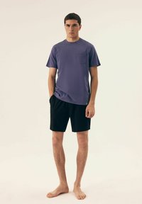 Fioletowa koszulka z krótkim rękawem z kieszonką na piersi, zestawiona z czarnymi szortami. Wykonana z bawełny, luźny krój i prosty design. Model boso.