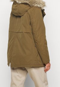 Brown hooded parka with faux fur trim. Features a straight cut, side pockets, and a water-resistant texture. Slightly longer back.