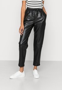Pantalons en simili cuir noir avec une taille élastique et un cordon de serrage, comprenant des poches latérales et un design de jambe effilée. Portés avec des baskets blanches.