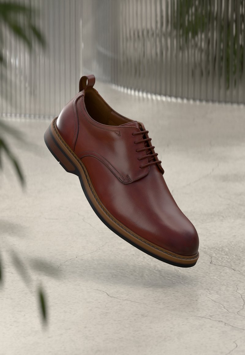 Brown leather lace-up dress shoe suspended above a light concrete floor with blurred plants and a corrugated glass wall in the background.