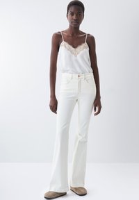 Jeans blancos acampanados combinados con una camiseta de seda ligera con un borde de encaje en el escote. La modelo está de pie frente a un fondo liso.