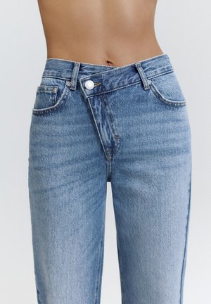 Középig érő kék farmernadrág aszimmetrikus átlapolt derékpánttal és egyetlen látható fehér gombbal, mezítelen hason viselve.