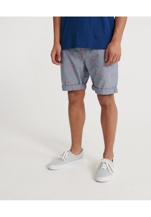 Homme portant un short bleu clair avec de petits motifs flamants roses roulés à l'ourlet, des baskets grises et une chemise bleu marine.