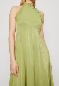 Woman wearing a sleeveless shiny light green dress with ruffled high neckline and fitted bodice, standing against a plain background.