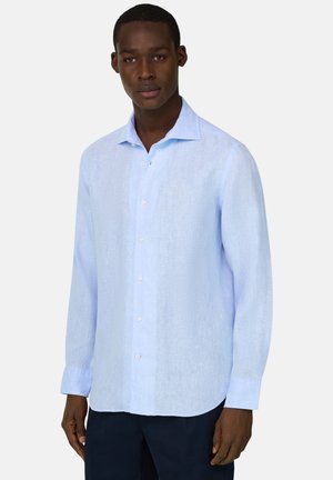 Homme noir portant une chemise boutonnée à manches longues bleu clair et un pantalon sombre, debout devant un fond blanc uni.