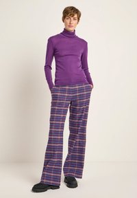Lila Rollkragenpullover kombiniert mit weit geschnittenen karierte Hosen in lila, blau und beige Tönen. Schwarze Schuhe mit dicker Sohle runden das Outfit ab.