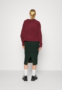 Pull en bordeaux à côtes avec un design court, associé à une jupe crayon vert foncé avec une fente au dos. Chaussures noires épaisses et chaussettes blanches.