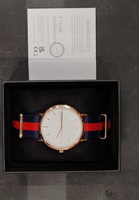 Gullfarget klokke med hvit urskive og marineblå-rød stripete nylonrem, vist i en svart eske med garantikort.