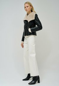 Giacca di pelle nera con colletto in pelliccia sintetica beige, chiusura con zip, abbinata a pantaloni bianchi a gamba larga e stivali neri con tacco a caviglia.