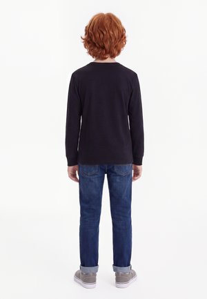 Barn med rødt krøllet hår står med ryggen mod kamera iført en sort langærmet trøje, blå bukser med opkrogede ben og grå sneakers på hvid baggrund.