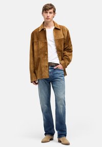 Brązowa zamszowa kurtka z kołnierzem, zapinana na guziki, z bocznymi kieszeniami, w połączeniu z jasnymi dżinsami i beżowymi butami.