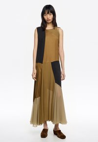 Sukienka maxi w brązowe i czarne bloki kolorów, z bezrękawową konstrukcją, warstwowym przezroczystym materiałem i zwiewnym dołem. Zamszowe brązowe buty.