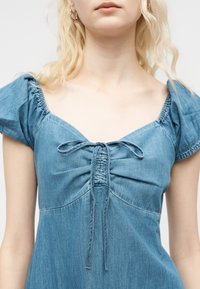 Personne blonde portant une robe en denim bleu avec des manches bouffantes et un devant froncé noué par un nœud au niveau de la poitrine.