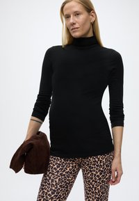 Γυναίκα που φοράει μαύρο ζιβάγκο, λεοπάρ κολάν, κρατώντας ένα σκούρο καφέ διπλωμένο μπουφάν, στέκεται μπροστά σε απλό λευκό φόντο.